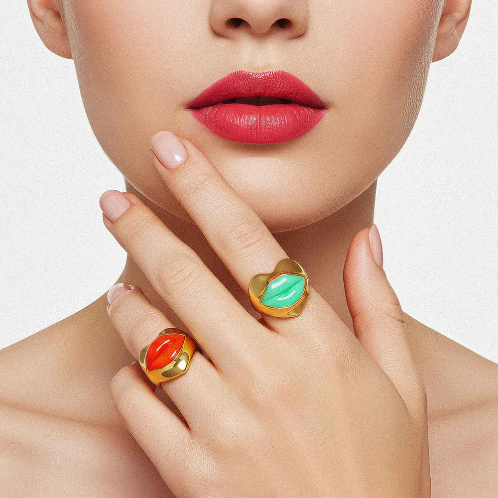 Close-up of a woman's face with red lipstick, hand with gold rings featuring green and orange stones, on a light background.
